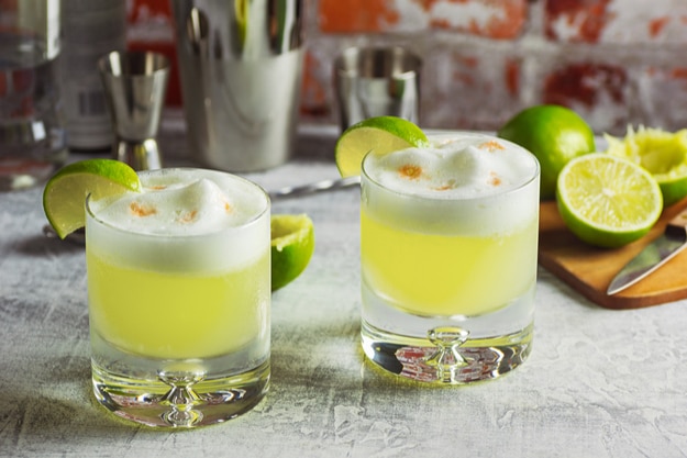 Two Pisco Sour Cocktails with Ingredients and Bottles on a Bar